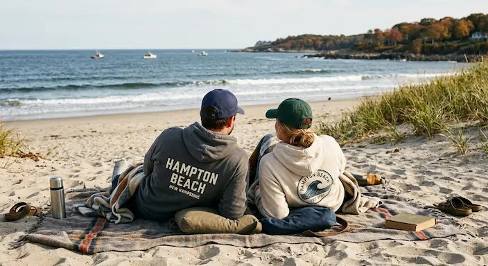 Two people sitting on a blanket on a sandy beach facing the ocean, wearing branded hoodies with Hampton Beach and Surf Life Boston logos on the back.