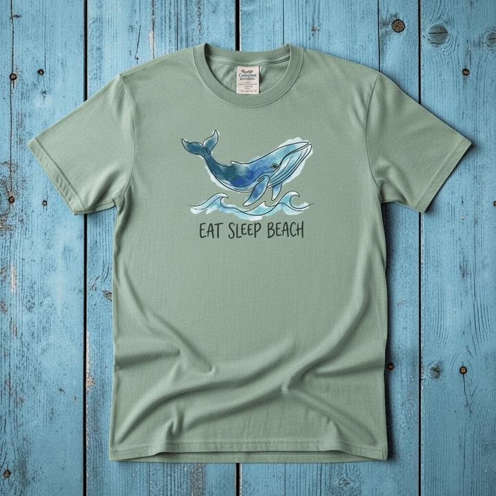 Flat lay - sage green Eat Sleep Beach tee with Comfort Colors label on weathered driftwood planks