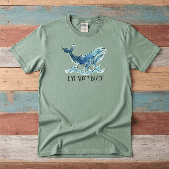 Flat lay - sage green Eat Sleep Beach tee with Comfort Colors label on weathered driftwood planks