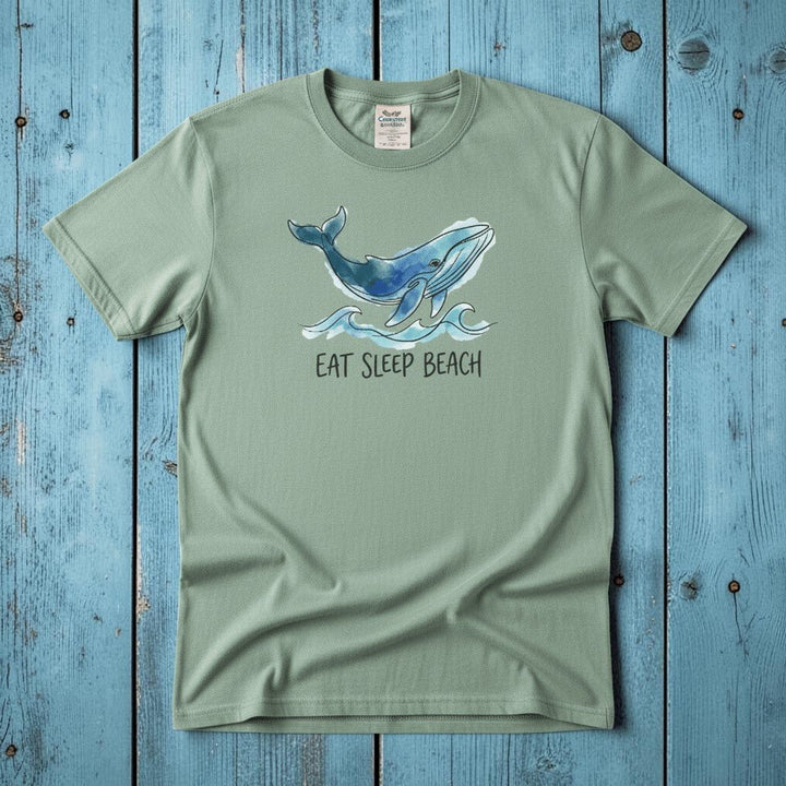 Flat lay - sage green Eat Sleep Beach tee with Comfort Colors label on weathered driftwood planks