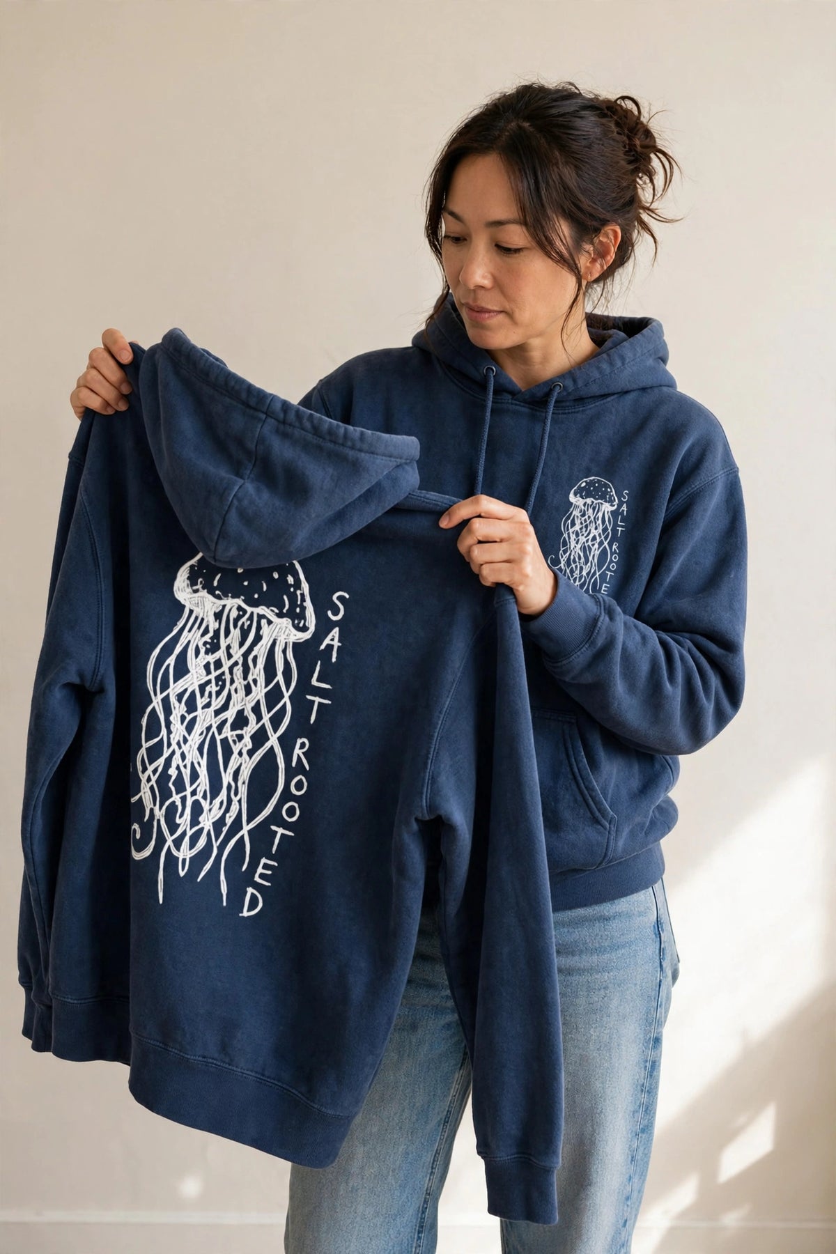 Woman wearing the Salt Rooted Jellyfish hoodie in True Navy, holding up a second hoodie showing the large back jellyfish print