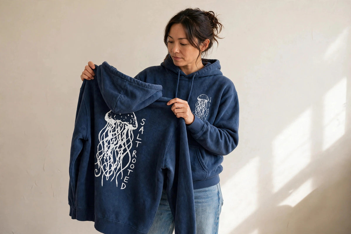 Woman wearing the Salt Rooted Jellyfish hoodie in True Navy, holding up a second hoodie showing the large back jellyfish print