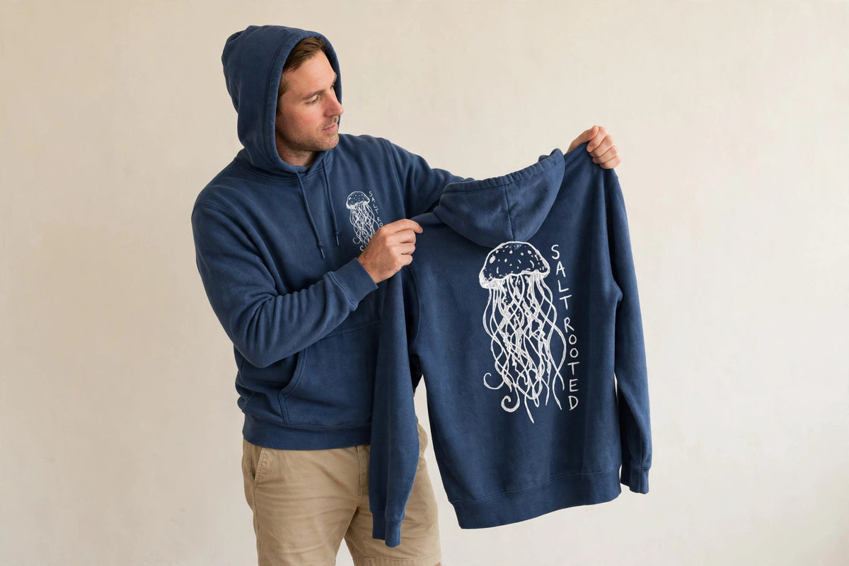 Man wearing the Salt Rooted Jellyfish hoodie in True Navy, holding up a second hoodie showing the large back jellyfish print