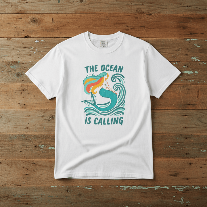 The Ocean Is Calling white garment-dyed mermaid wave tee laid flat on rustic reclaimed wood