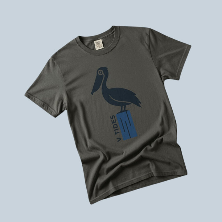 Pelican Dock V Tides coastal graphic t-shirt in Pepper dark charcoal casually tossed on a light sand-colored woven rug, coastal lifestyle product photo