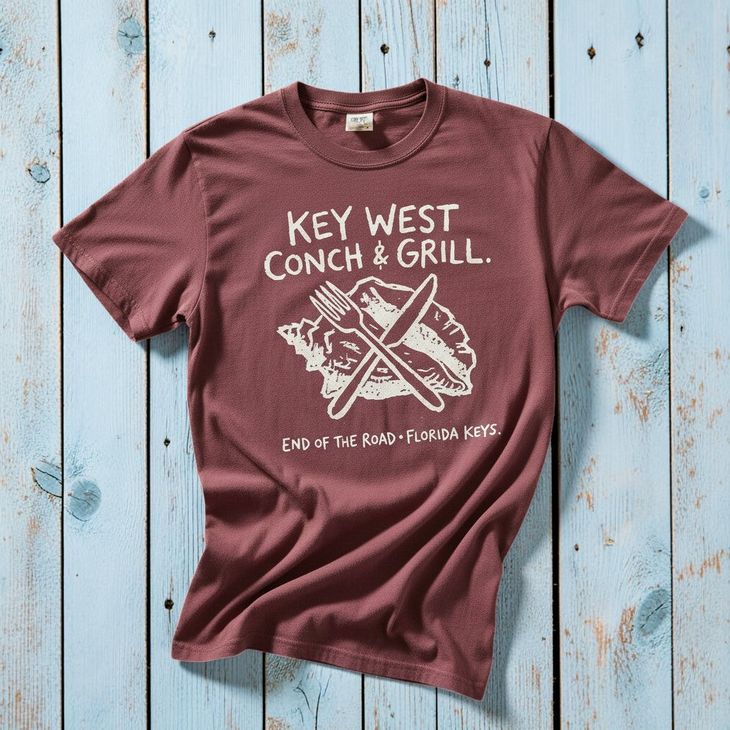 Key West Conch and Grill graphic t-shirt in Brick mauve with cream graphic casually tossed on a light sand-colored woven rug, Florida Keys lifestyle product photo