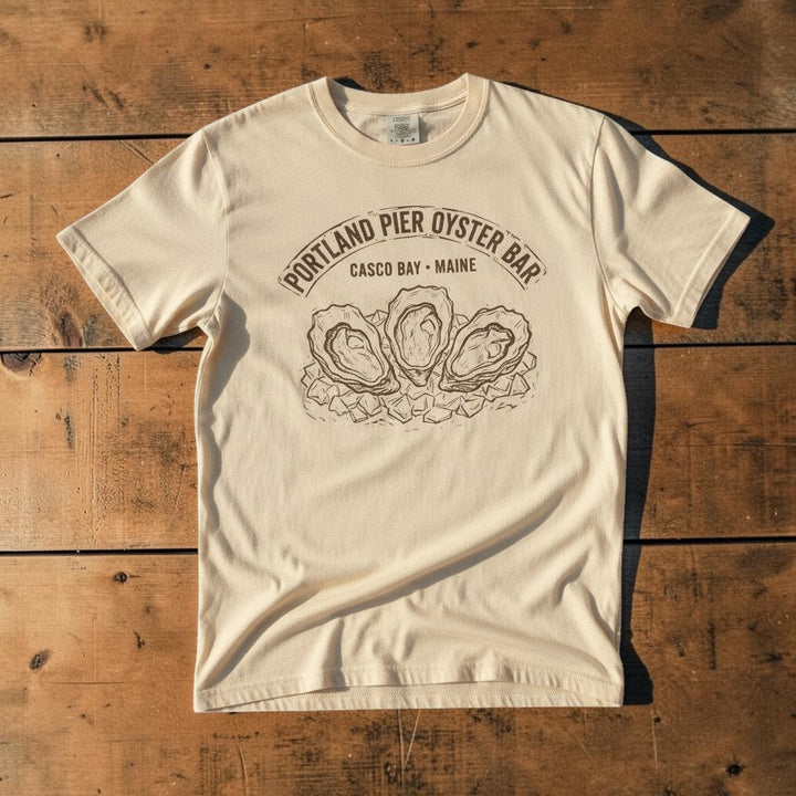 Portland Pier Oyster Bar Casco Bay Maine vintage seafood graphic t-shirt in Ivory casually tossed on a light sand-colored woven rug, coastal lifestyle product photo
