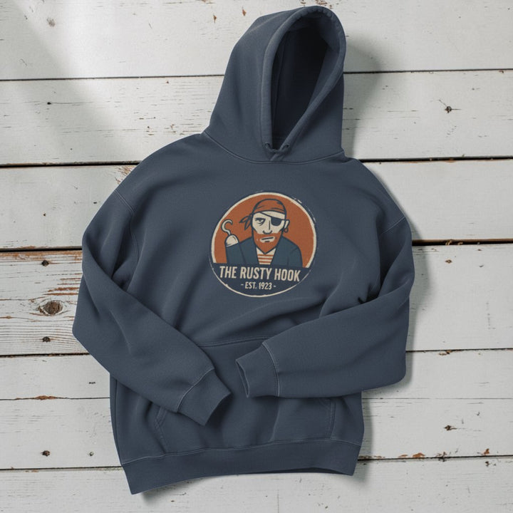 The Rusty Hook Hoodie in Steel Blue — front pirate badge graphic, warm sand studio background