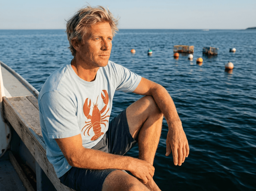 Man sitting on a dock by the water wearing a t-shirt with a lobster graphic.