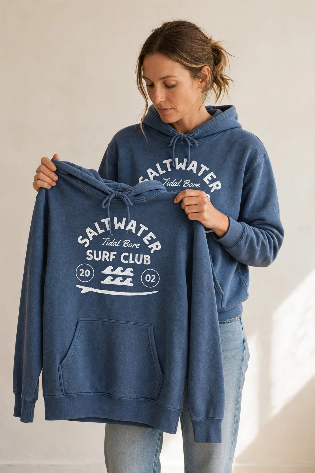 Woman wearing the Saltwater Tidal Bore Surf Club Hoodie in Denim, holding up the front print