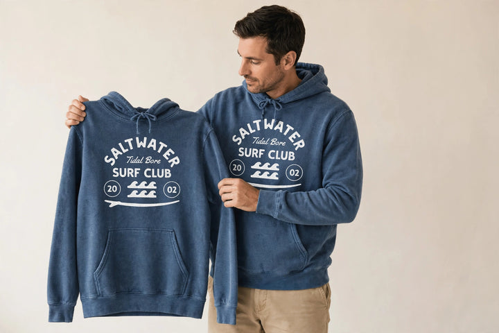 Man wearing the Saltwater Tidal Bore Surf Club Hoodie in Denim, holding up the front print