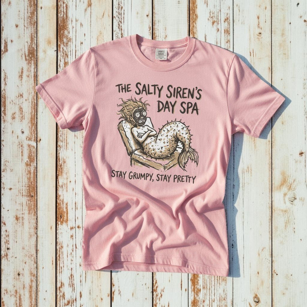 The Salty Siren's Day Spa mermaid graphic t-shirt in Blossom pink casually tossed on a light sand-colored woven rug